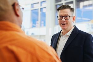 Man in suit and glasses talking to man in orange dress shirt in an office setting